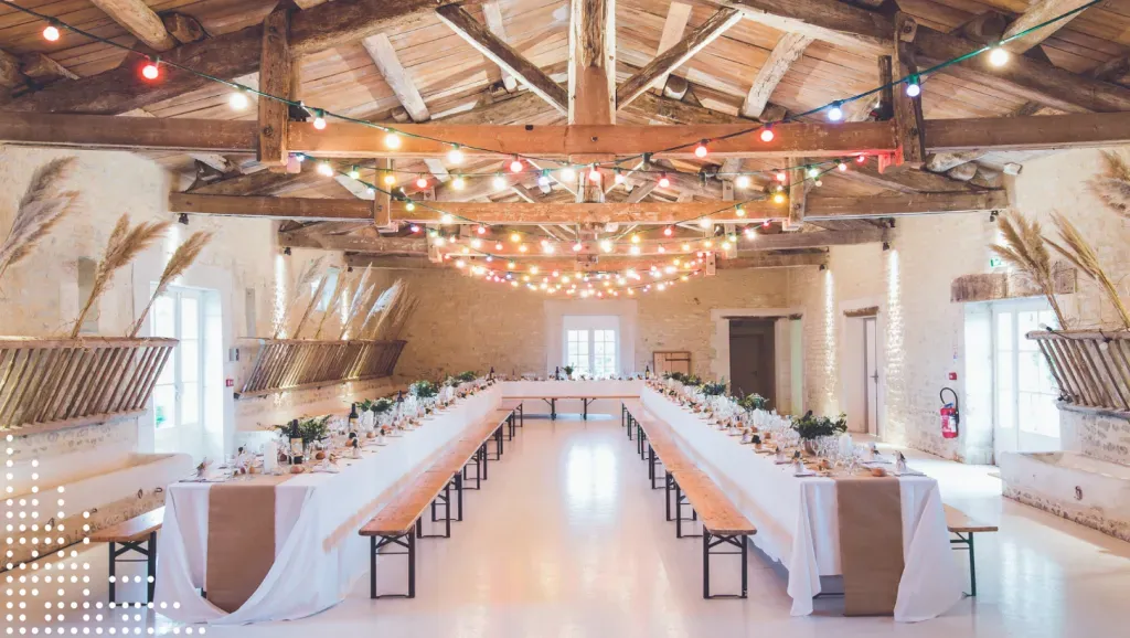 Rustic indoor wedding reception setup with long banquet tables, wooden beams, and colorful string lights.