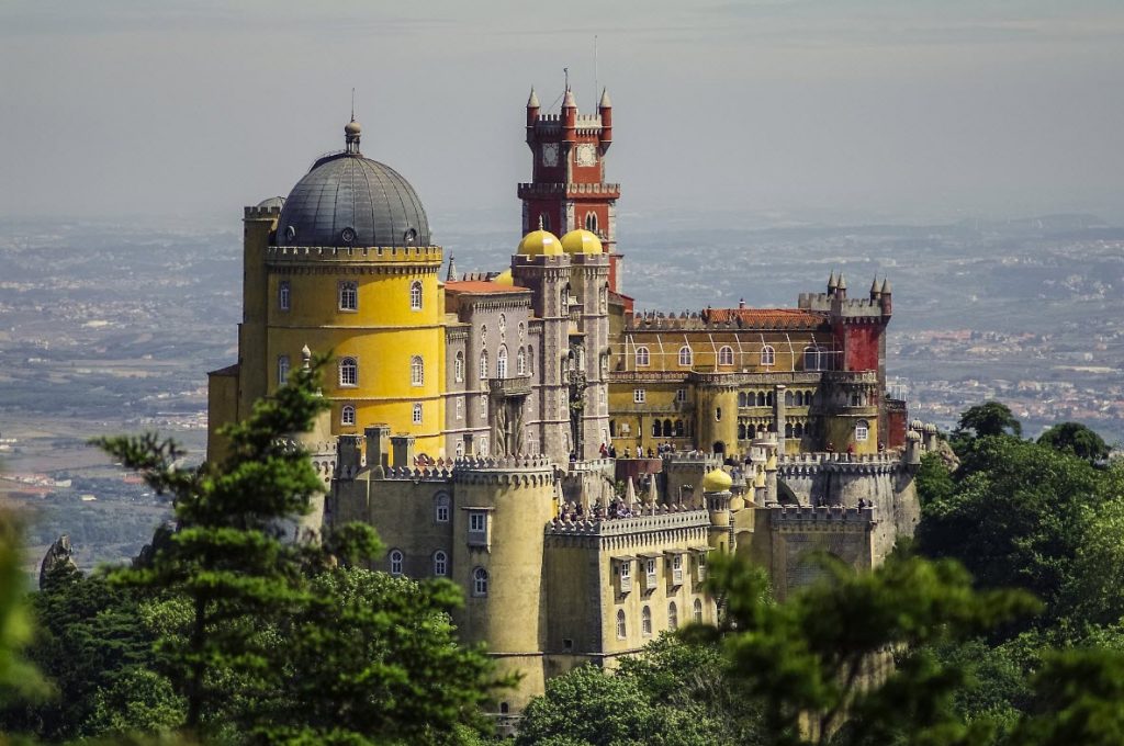 Yellow and red palace with domes and turrets, surrounded by greenery, overlooking a town.