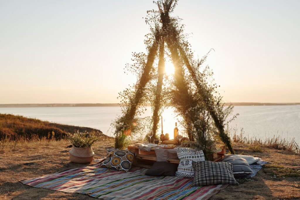 Picnic setup with pillows and a blanket by a lakeside at sunset.