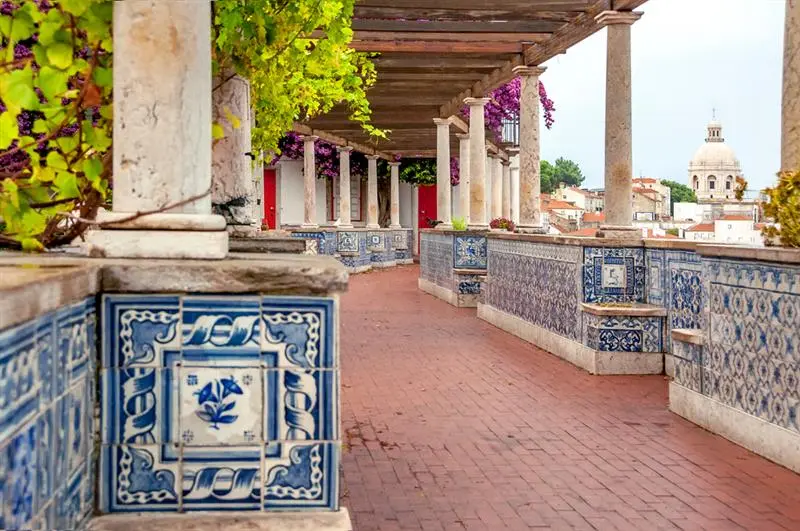 A picturesque corridor adorned with blue tiles, wooden beams, and vibrant foliage, leading to a scenic view of distant buildings.