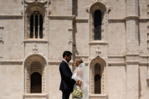 Bride and groom standing in profile outside the detailed stone facade of Jerónimos Monastery in Lisbon on a sunny day