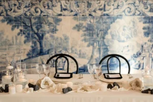 An elegant dining setup featuring two black chairs, crystal glassware, candles, and natural stone decorations against a blue-tiled wall.