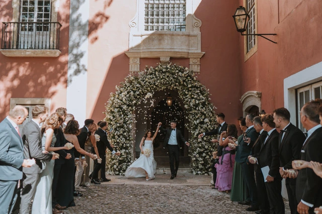 A joyful couple exits a floral-decorated archway, celebrating their wedding as guests cheer and toss petals in a picturesque setting.