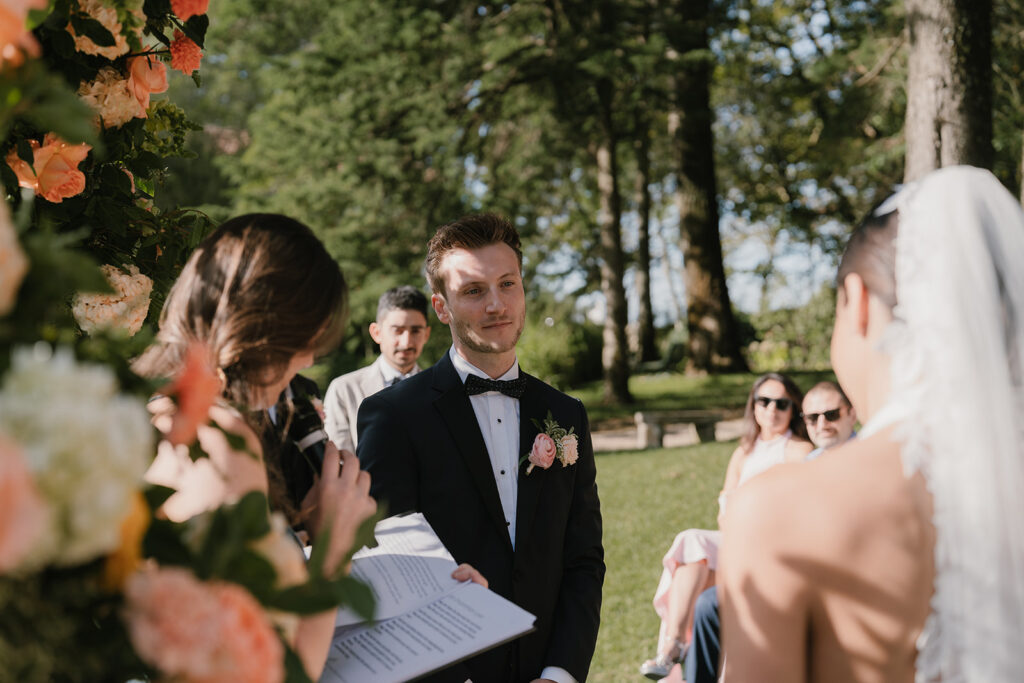 The groom, in a black tuxedo and bowtie, looks toward his bride during an outdoor ceremony, framed by flowers in the foreground.