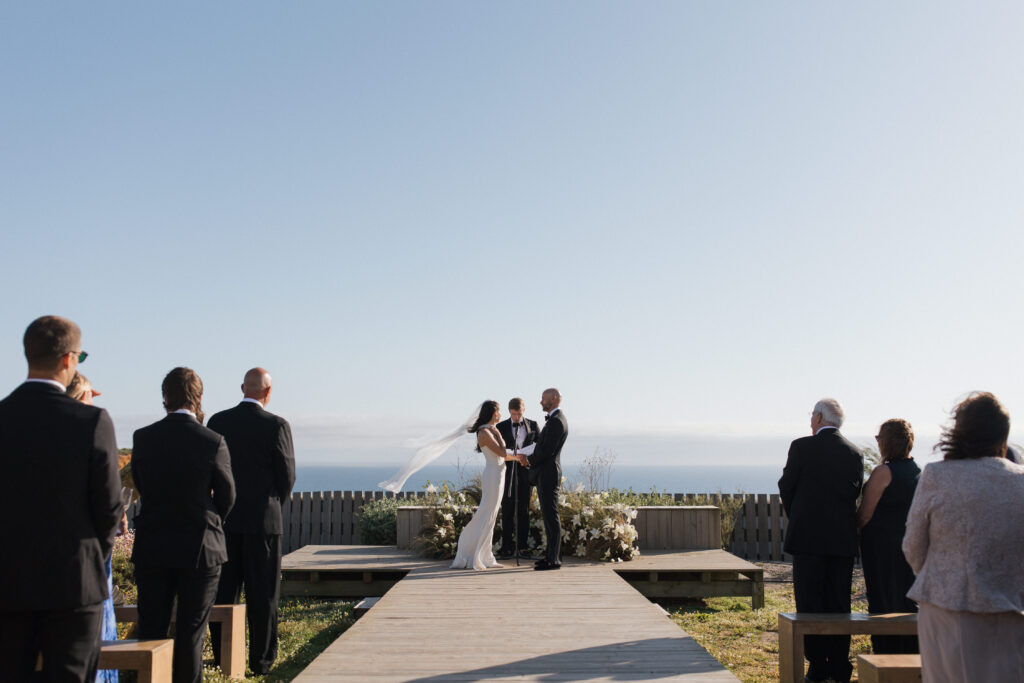 A wide shot of an outdoor wedding ceremony where the couple, facing each other, stands on a wooden platform overlooking the ocean under a vast blue sky.