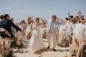Joyful bride and groom walking down a sunny beach aisle decorated with pampas grass as guests cheer and throw petals