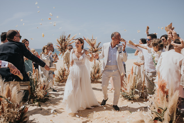Joyful bride and groom walking down a sunny beach aisle decorated with pampas grass as guests cheer and throw petals