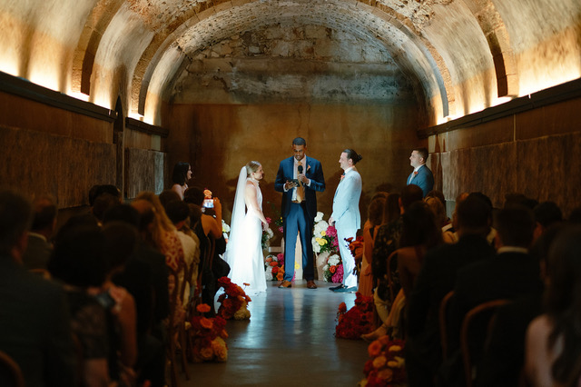 A wedding ceremony taking place in a dimly lit, arched stone or brick venue, with the couple, an officiant, and guests visible.