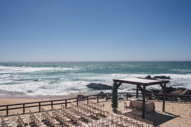 Beachfront wedding venue with rows of empty chairs overlooking the ocean, under a clear blue sky and a wooden pergola.