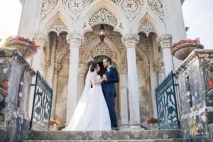 A bride in a flowing white dress and a groom in a blue suit share a loving gaze at an ornate stone entrance, surrounded by floral accents.