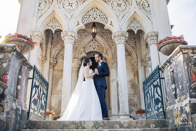 A bride in a flowing white dress and a groom in a blue suit share a loving gaze at an ornate stone entrance, surrounded by floral accents.