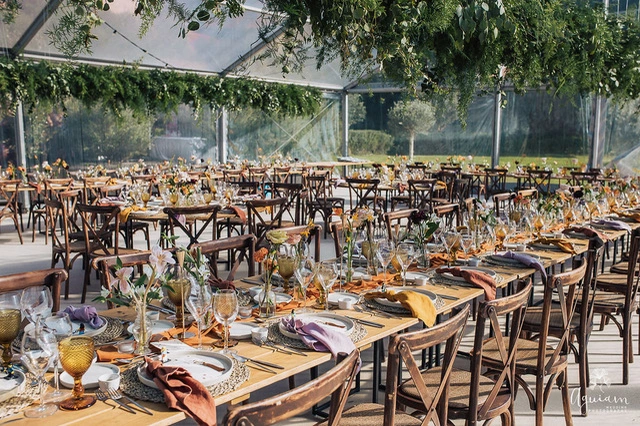 Elegantly set dining tables with colorful napkins and glassware, under a transparent tent adorned with greenery, ready for an event.