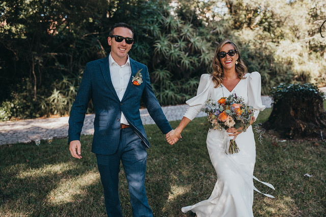 A newly married couple in sunglasses walking hand-in-hand through a sunny, leafy park or garden.