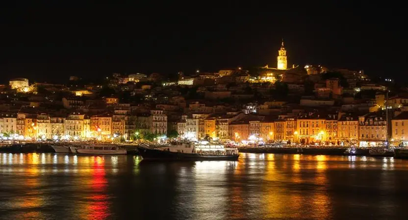 Illuminated harbor at night, showcasing a hillside town with colorful buildings and a tower, reflecting in calm waters.