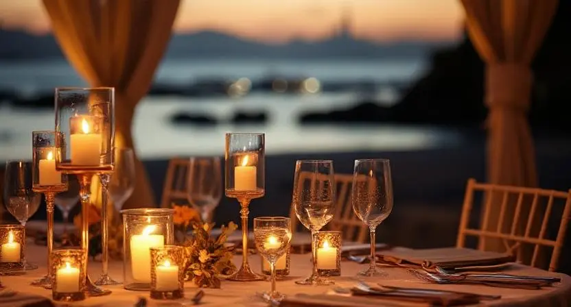 Elegant candlelit dinner setup on the beach at sunset, featuring glassware and floral accents amid a serene coastal backdrop.