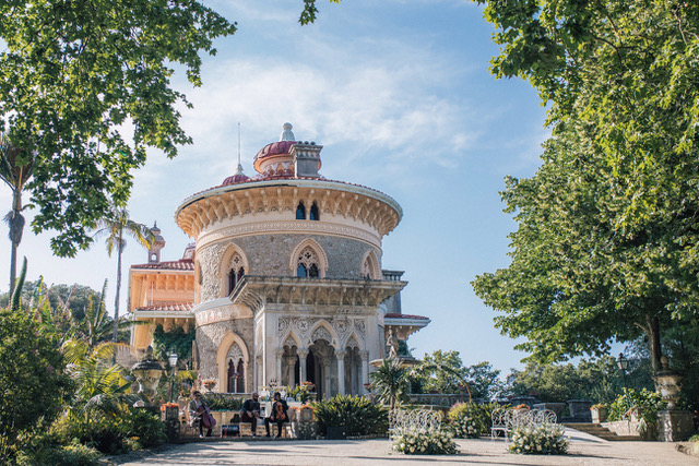 An ornate, round stone palace or villa with a red roof, set in a sunny garden and framed by large green trees.
