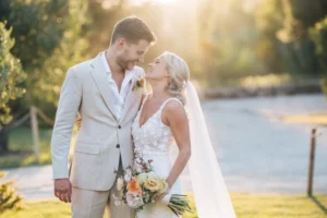 A couple shares a tender moment outdoors during sunset, surrounded by greenery, with the bride holding a bouquet of flowers.