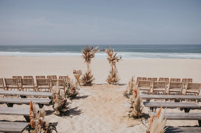 A serene beach wedding setup with wooden benches, floral decorations, and an ocean backdrop under a clear blue sky.