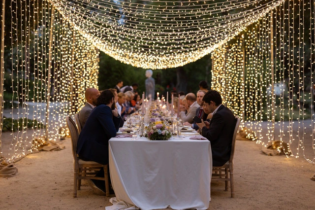 A beautifully arranged outdoor dinner table adorned with candles and flowers, illuminated by sparkling fairy lights overhead.