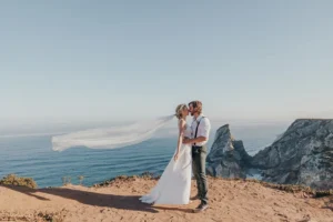 A couple shares a kiss on a cliff overlooking the ocean, with a flowing wedding veil and stunning coastal scenery in the background.