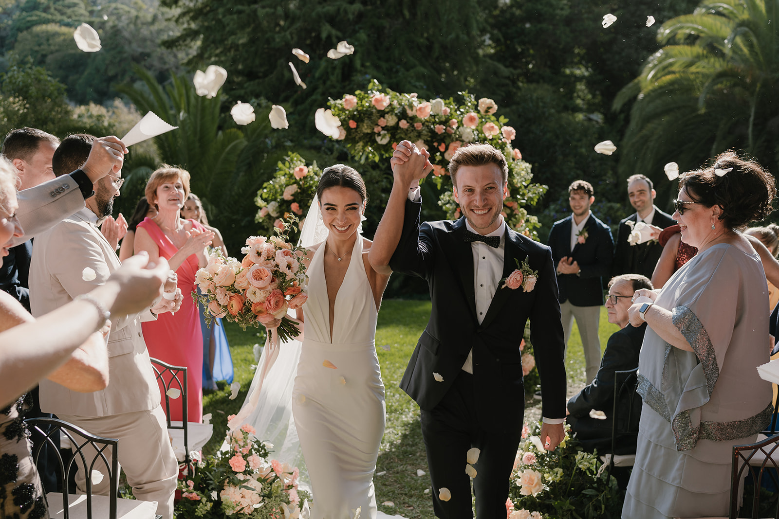 A smiling newlywed couple walks down an aisle lined with pink and orange roses as guests clap and toss white petals.