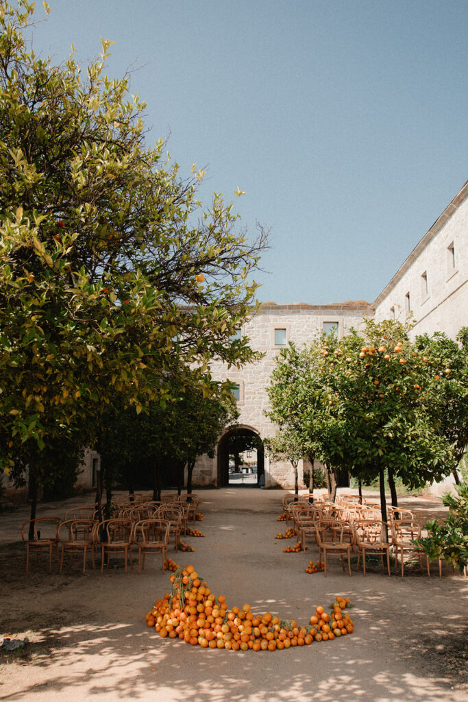 An outdoor wedding ceremony setup in a courtyard with rows of wooden chairs, orange trees, and an arch of oranges in the foreground.