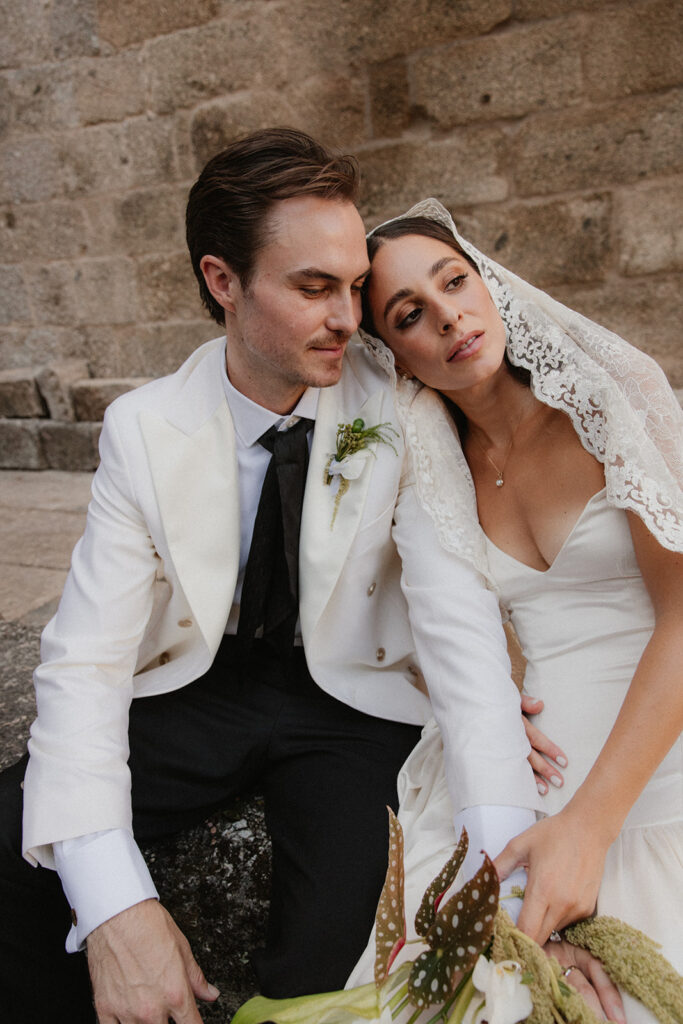 A groom in a white jacket and black pants sits with his bride, who wears a simple white dress and lace veil, as she rests her head on his shoulder.