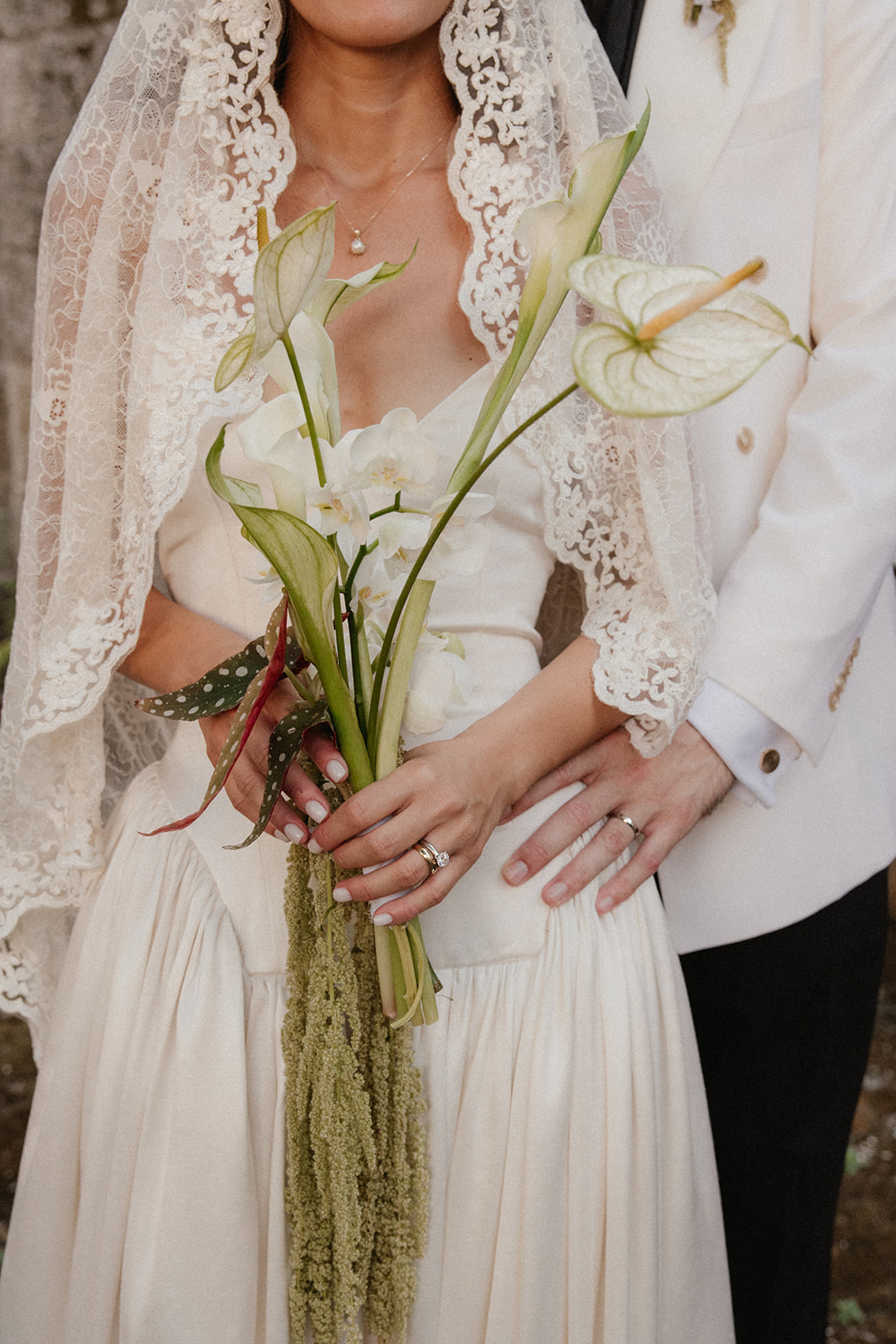 A close-up of a bride holding a bouquet of white calla lilies and green hanging amaranthus, with the groom's arm around her waist.