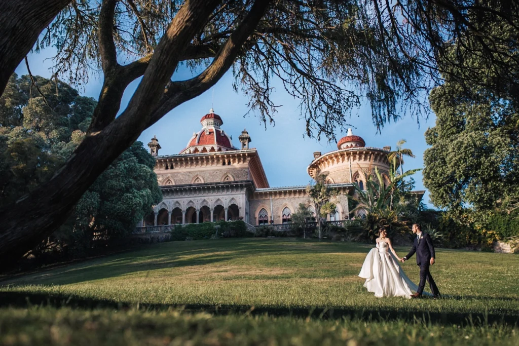 A newly married couple in a white dress and black suit walks hand-in-hand across a grassy lawn towards a grand, ornate palace with a red roof, framed by a large dark tree.