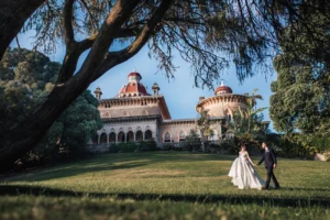 A newly married couple in a white dress and black suit walks hand-in-hand across a grassy lawn towards a grand, ornate palace with a red roof, framed by a large dark tree.